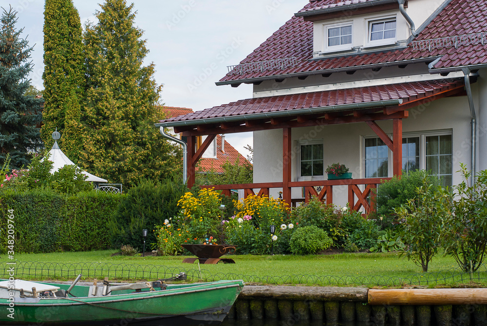 Beautiful white house with red roof on the river bank. In front of it there is a green lawn and a garden, a green boat is parked by the bank.