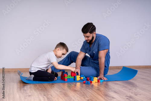 Young male doctor with beard in a blue uniform sitting on the floor next to the boy 10 years, they play educational toys, concept rehabilitation