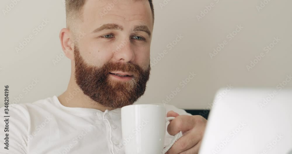 Close up view of concentrated man drinking hot coffee with rich aroma while working in bed. Handsome bearded guy in 30s sitting on bed while using his laptop. Concept of freelance work. Close up view of concentrated man drinking hot coffee with rich aroma while working in bed. Handsome bearded guy in 30s sitting on bed while using his laptop. Concept of freelance work.