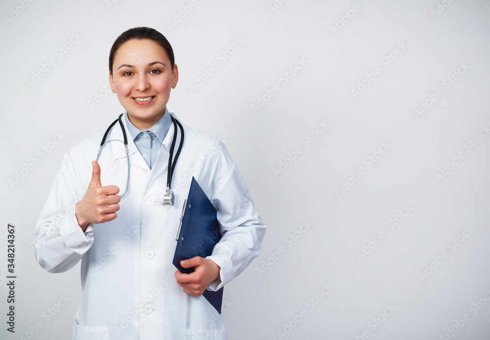 Smiling young doctor woman with stethoscope showing thumbs up isolated on white background. Female doctor in medical gown holding health card on notepad folder. Smiling young doctor woman with stethoscope showing thumbs up isolated on white background. Female doctor in medical gown holding health card on notepad folder.