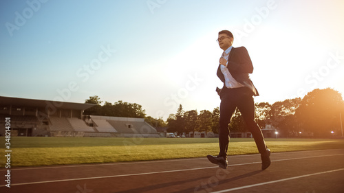 Young Serious Businessman in a Suit Running in an Outdoors Stadium. He Wears Glasses and is Holding a Mobile Phone. Office Worker Chasing Goals. Management Satire.