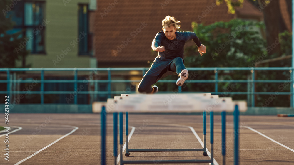 Fototapeta premium Athletic Fit Man in Grey Shirt and Shorts Hurdling in the Stadium. He is Jumping Over Barriers on a Warm Summer Afternoon. Athlete Doing His Routine Sports Practice.