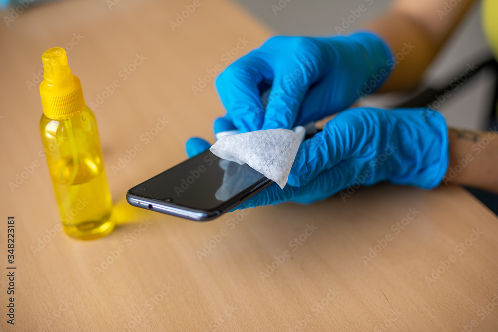 Woman's hand in blue gloves sanitizing cleaning smartphone mobile phone with alcohol on wood table surface with wet wipes