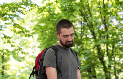 A young Caucasian man with a beard and a backpack, stands among the forest with a displeased and angry face.