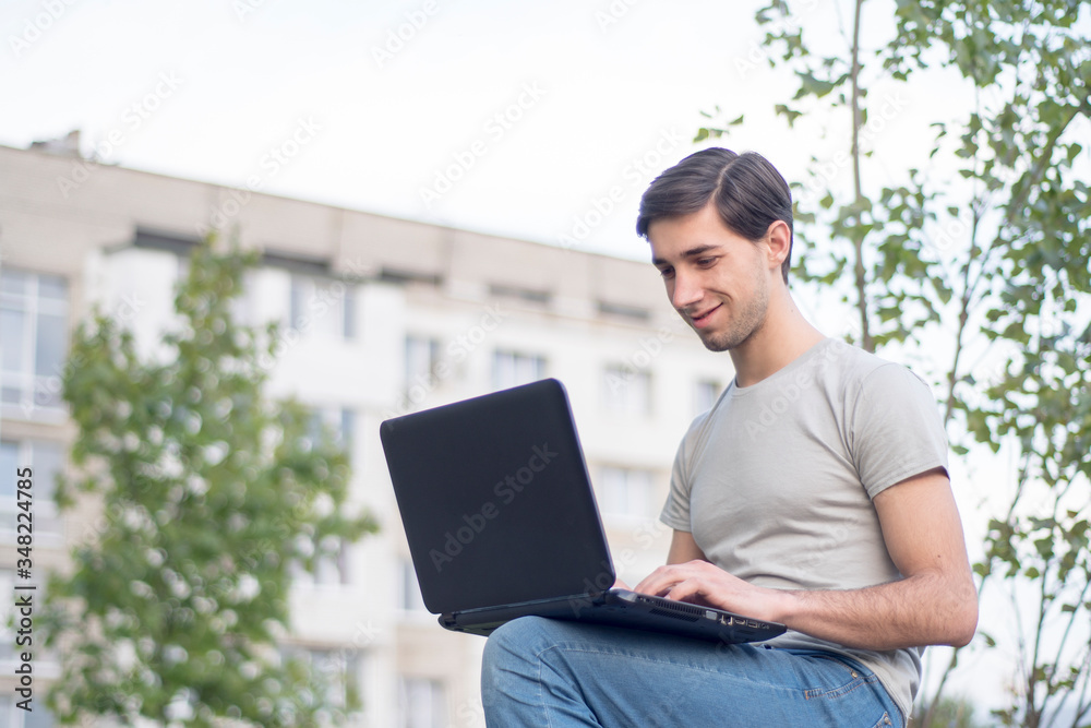 A young caucasian man sitting in a park, smiling and typing on a laptop. Remote work