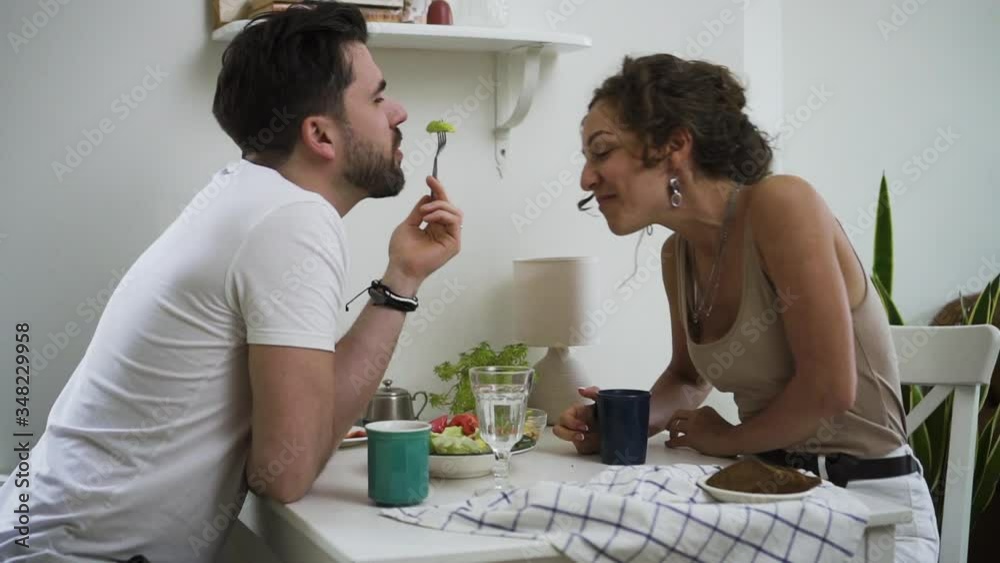 Bearded man and beautiful woman are eating dinner and kissing at table in home kitchen spbd. Young happy couple eats tasty food and kisses with tenderness, sitting at desk in modern interior. Two Bearded man and beautiful woman are eating dinner and kissing at table in home kitchen spbd. Young happy couple eats tasty food and kisses with tenderness, sitting at desk in modern interior. Two
