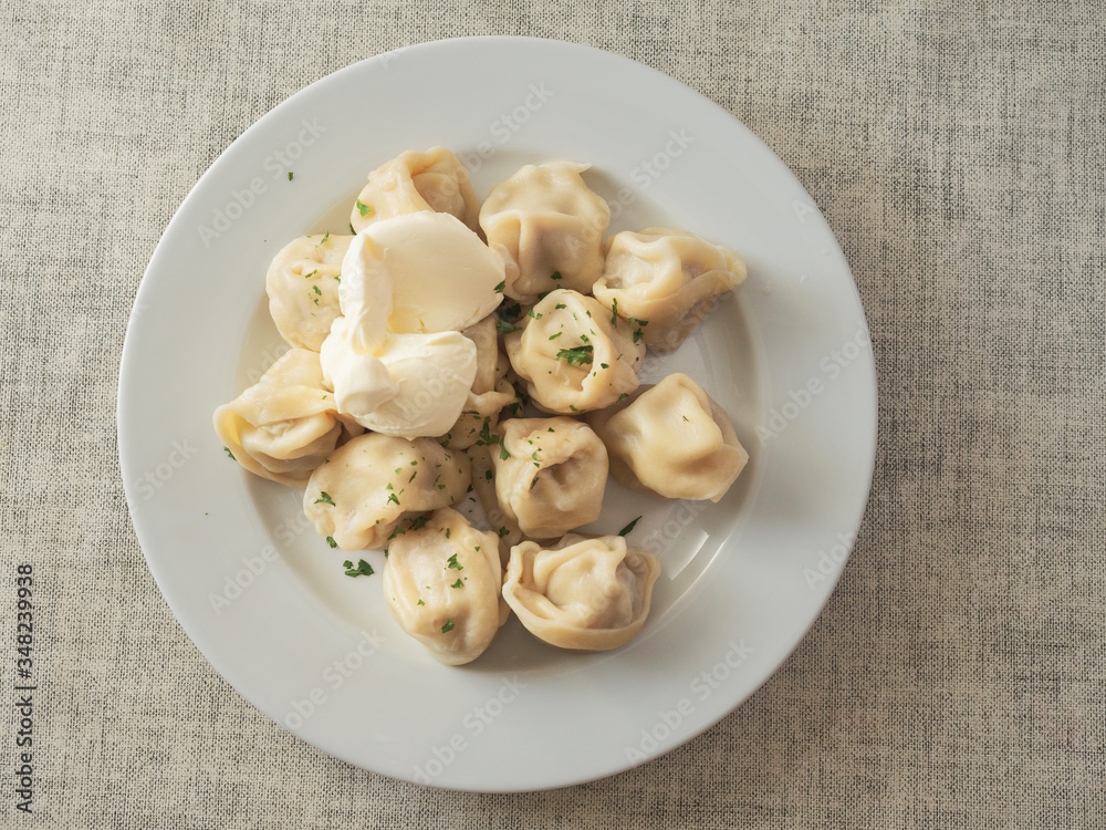Serving of russian traditional dumplings on white plate with sour cream, top view. Plate on simple white table clothe. Serving of russian traditional dumplings on white plate with sour cream, top view. Plate on simple white table clothe.