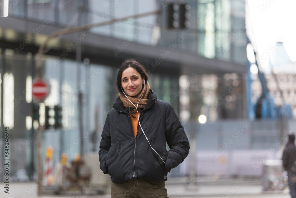 Fototapeta premium Young Indian woman wearing jacket and listening to earphones on street smiling sincerely at camera.