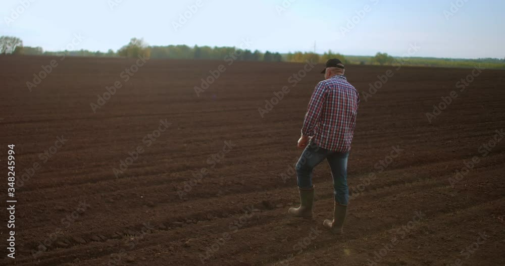 Farmer walking in idyllic,rural plowed field at sunset. Farmer wiping off sweat from forehead while walking on the field at sunset. Rubber boots for work use. A worker go with rubber boots at sunset Farmer walking in idyllic,rural plowed field at sunset. Farmer wiping off sweat from forehead while walking on the field at sunset. Rubber boots for work use. A worker go with rubber boots at sunset
