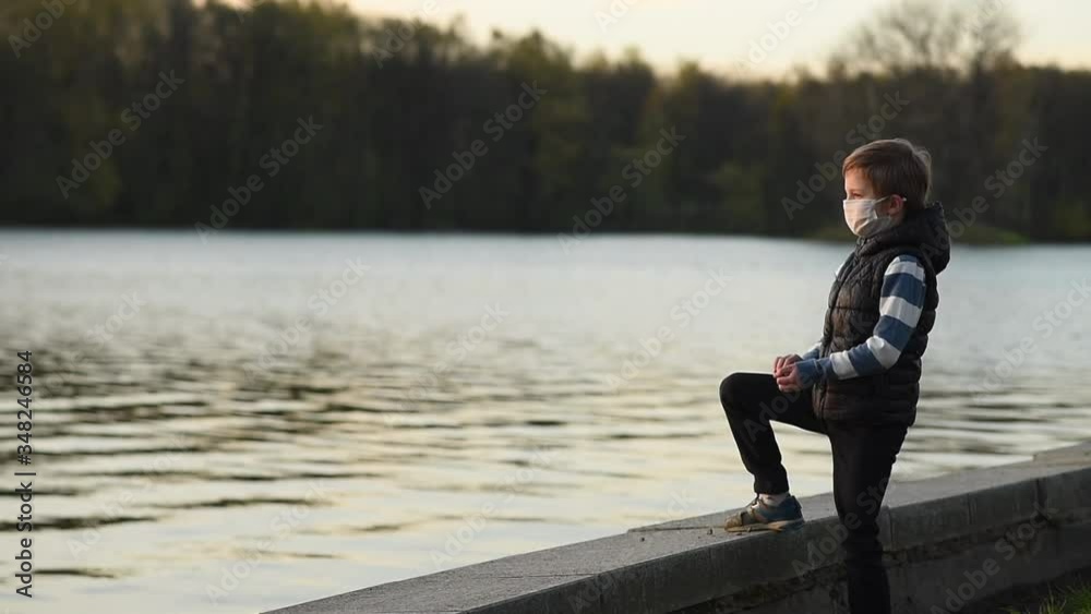 A boy in a medical mask on the shore throws stones into the water at sunset in slow mo A boy in a medical mask on the shore throws stones into the water at sunset in slow mo