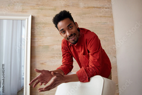 Glad young brown-eyed dark skinned brunette guy with short haircut looking positively at camera while leaning on chair, standing over home interior in red shirt