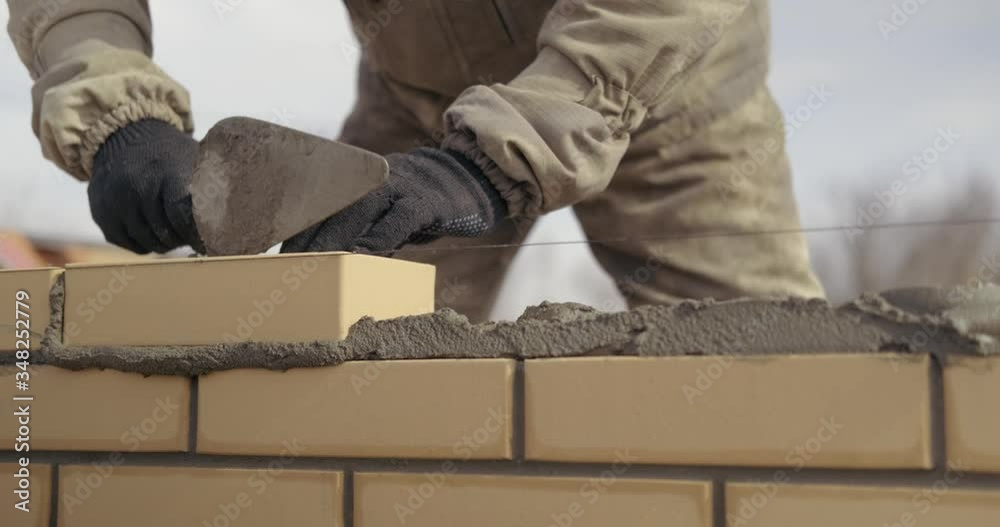 Bricklayer works at construction site, works as trowel, puts kippichi, builds wall of house, works with level, taps masonry with trowel, smoothes brick, removes excess mortar, even brickwork Bricklayer works at construction site, works as trowel, puts kippichi, builds wall of house, works with level, taps masonry with trowel, smoothes brick, removes excess mortar, even brickwork