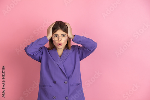 Shocked holding head. Caucasian young woman's portrait isolated on pink studio background. Beautiful female model in purple jacket. Concept of human emotions, facial expression, sales, ad. Copyspace. © master1305