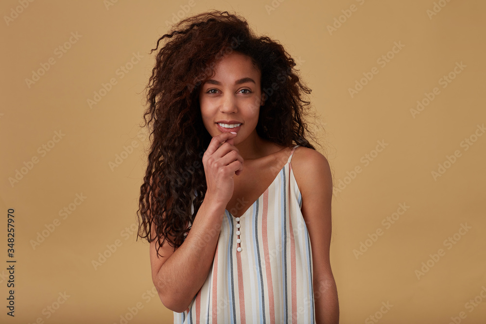 Glad young attractive curly dark skinned brunette woman with natural makeup touching gently her face and looking cheerfully at camera, isolated over beige background Glad young attractive curly dark skinned brunette woman with natural makeup touching gently her face and looking cheerfully at camera, isolated over beige background