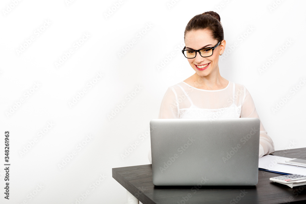 Smiling and successful woman wearing blouse and eyeglasses in sits at her desk and looking at a laptop. The concept successful and productive manager. White background. Copy paste Smiling and successful woman wearing blouse and eyeglasses in sits at her desk and looking at a laptop. The concept successful and productive manager. White background. Copy paste