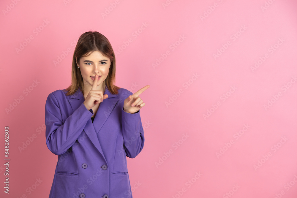 Pointing secretly. Caucasian young woman's portrait isolated on pink studio background. Beautiful female model in purple jacket. Concept of human emotions, facial expression, sales, ad. Copyspace. Pointing secretly. Caucasian young woman's portrait isolated on pink studio background. Beautiful female model in purple jacket. Concept of human emotions, facial expression, sales, ad. Copyspace.