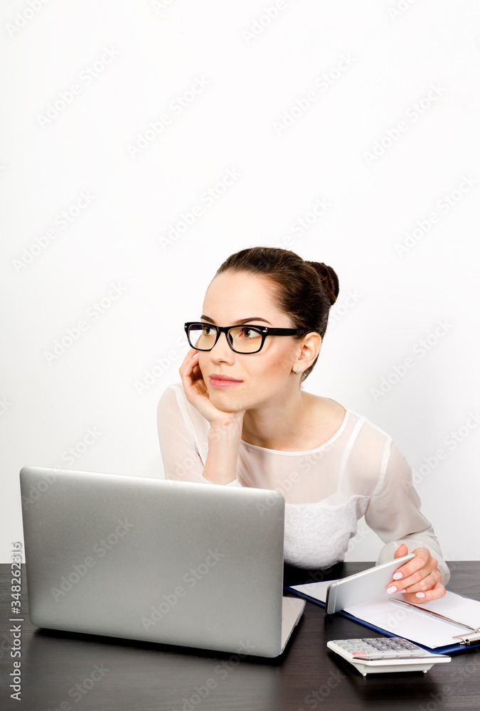 young successful woman entrepreneur wearing eyeglasses sits at her desk on a white background at her laptop, holds her mobile phone and looks away thoughtfully. young successful woman entrepreneur wearing eyeglasses sits at her desk on a white background at her laptop, holds her mobile phone and looks away thoughtfully.