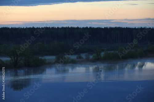 An image of a blue morning on a lake with a clear sky on the horizon, the reflection of a forest in the water in a creeping fog, and a single buoy on the surface.Peaceful landscape at dusk.Russia