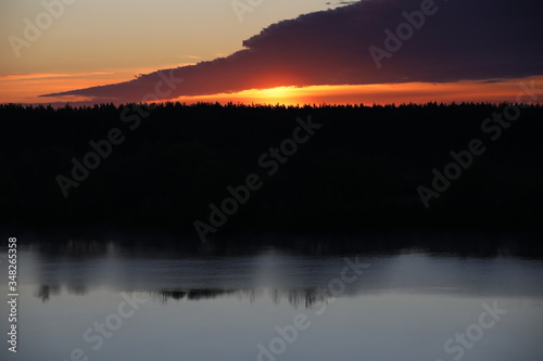 Dramatic sky background.The Golden light of the rising sun at dusk illuminates the overhanging continuous cloud on the clear horizon and the forest reflected in the ripples of a calm lake.Russia © Nina