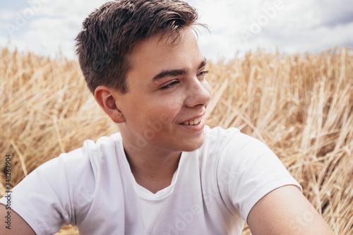 Close-up portrait of cheerful handsome teenage boy, sitting on wheat field, under a sky with clouds, at summer day.