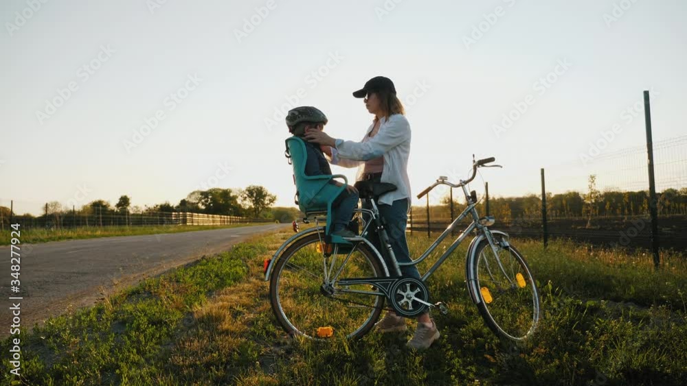 Cycling. The child is sad, mom puts a bicycle helmet on the head of her little son by standing on the roadside. Child should wear bicycle helmet Cycling. The child is sad, mom puts a bicycle helmet on the head of her little son by standing on the roadside. Child should wear bicycle helmet