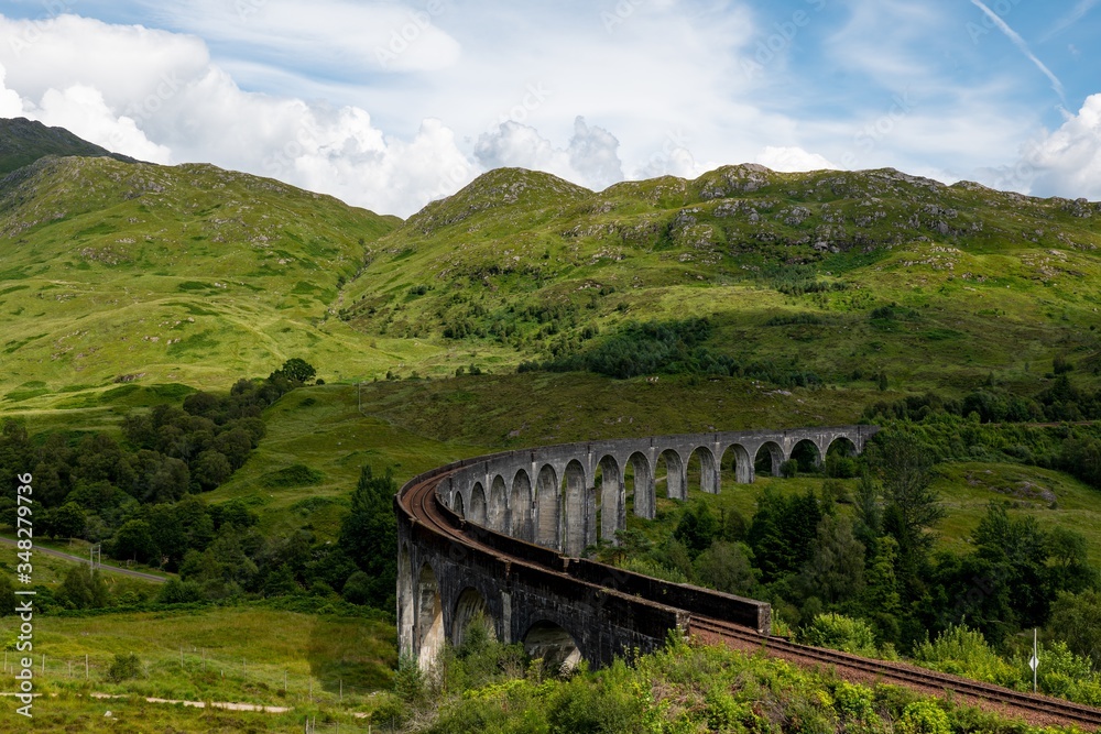 The landscape with famous Glenfinnan Viaduct in Scotland with a historic steam train in the background with cloud shadows