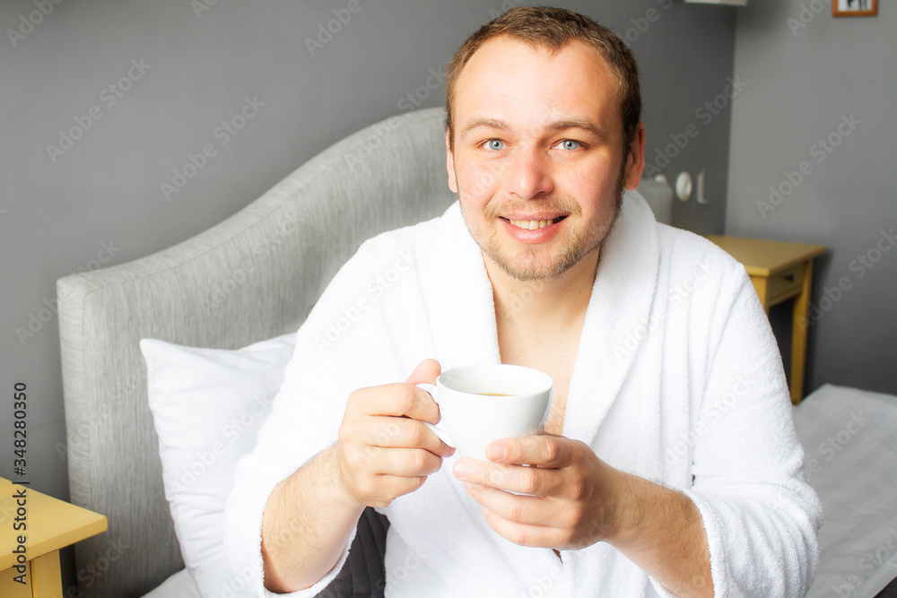 Portrait of a young man in a bathrobe with a cup of coffee at home. Handsome man is drinking coffee in a hotel bed. Portrait of a young man in a bathrobe with a cup of coffee at home. Handsome man is drinking coffee in a hotel bed.