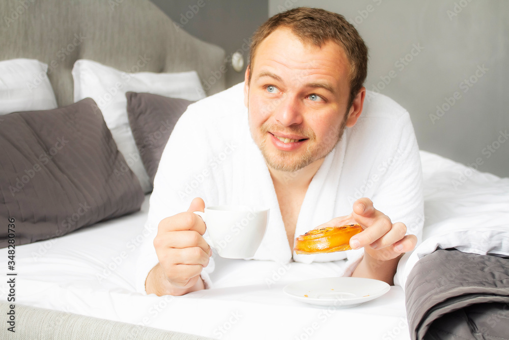 Handsome young man having breakfast while lying in bed. The guy on the white bed with food. Breakfast in bed. Handsome young man having breakfast while lying in bed. The guy on the white bed with food. Breakfast in bed.