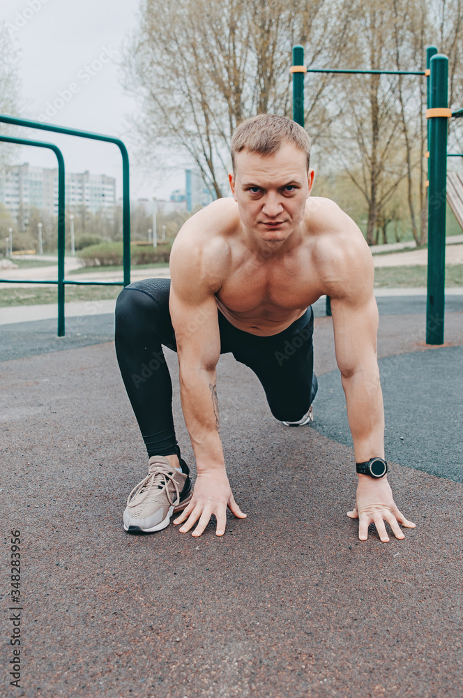 Obraz premium A young guy stretches and warms up on the sports ground. Athlete man in running pose on city street. Sport tight clothes. Bright sunset. sport man on starting position 20-30s