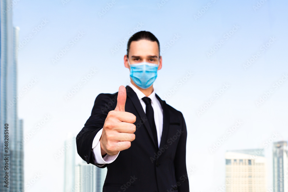 Thumbs up! Young businessman giving approval hand gesture towards camera and wearing blue anti virus medical mask. Business male agent portrait against modern city and blue sky background. Thumbs up! Young businessman giving approval hand gesture towards camera and wearing blue anti virus medical mask. Business male agent portrait against modern city and blue sky background.