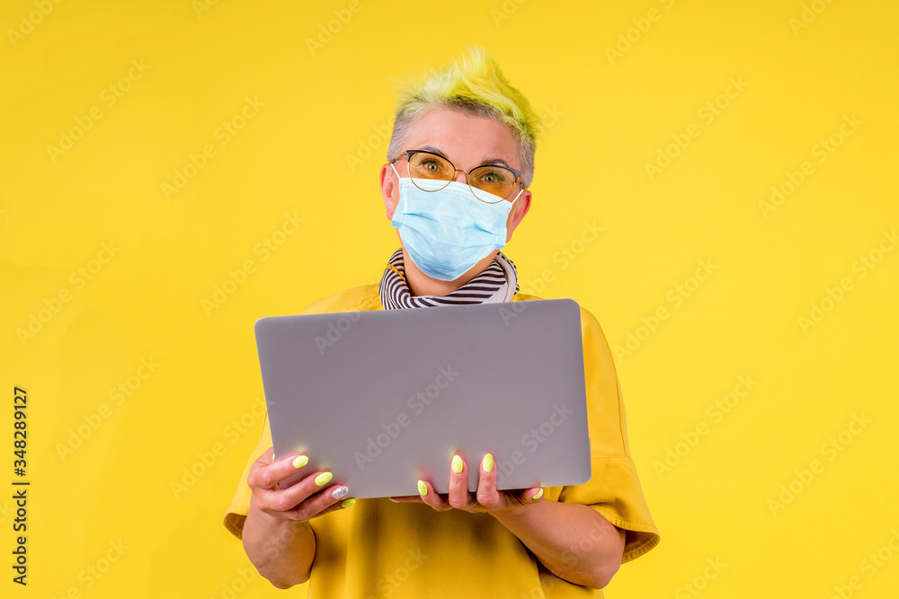 mature old woman with colored stylish yellow dyed hair in warm dress and medical mask holding modern laptop background studio