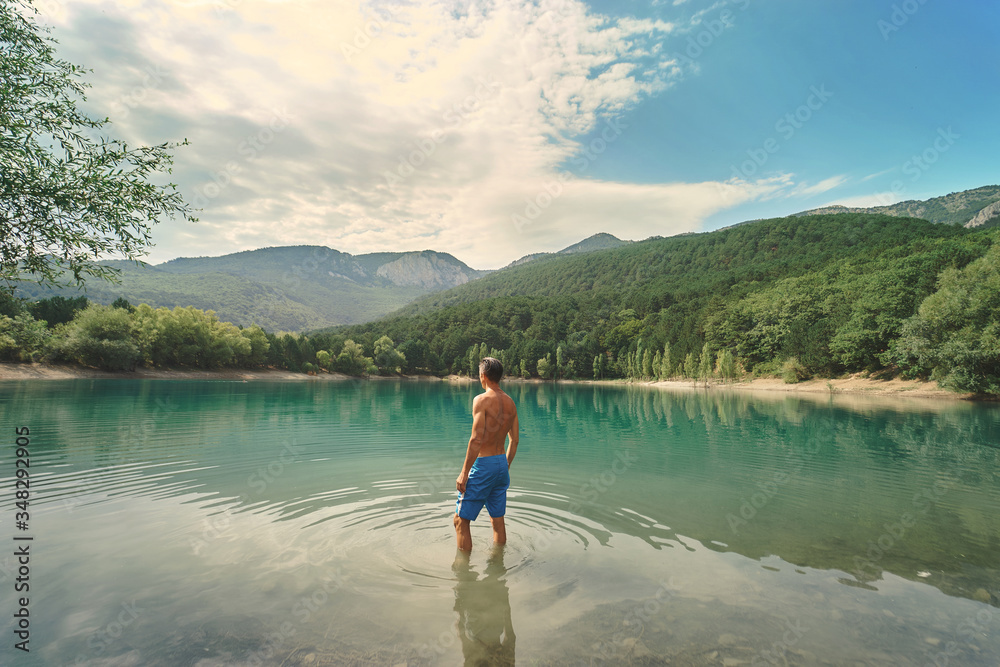 back view athletic man in blue swimming trunks standing by clear blue mountain lake, enjoying beautiful nature scenery. people in nature and travel concept