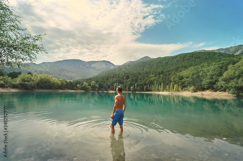 back view athletic man in blue swimming trunks standing by clear blue mountain lake, enjoying beautiful nature scenery. people in nature and travel concept
