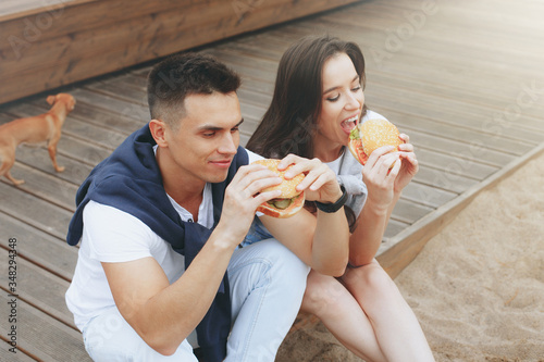 Young beautiful positive couple eating hambergers sitting on beach photo