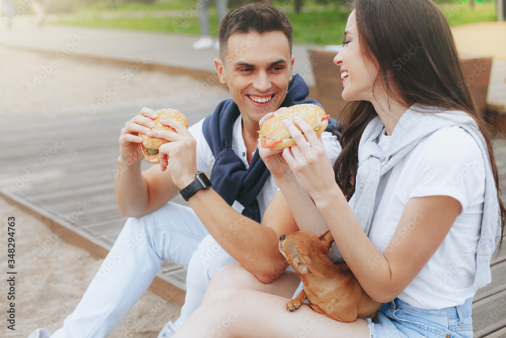 Young beautiful positive couple eating hambergers sitting on beach