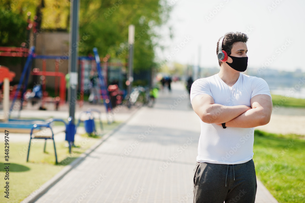 Portrait sports arabian man in black medical face mask posed outdoor with earphones during coronavirus quarantine. Portrait sports arabian man in black medical face mask posed outdoor with earphones during coronavirus quarantine.