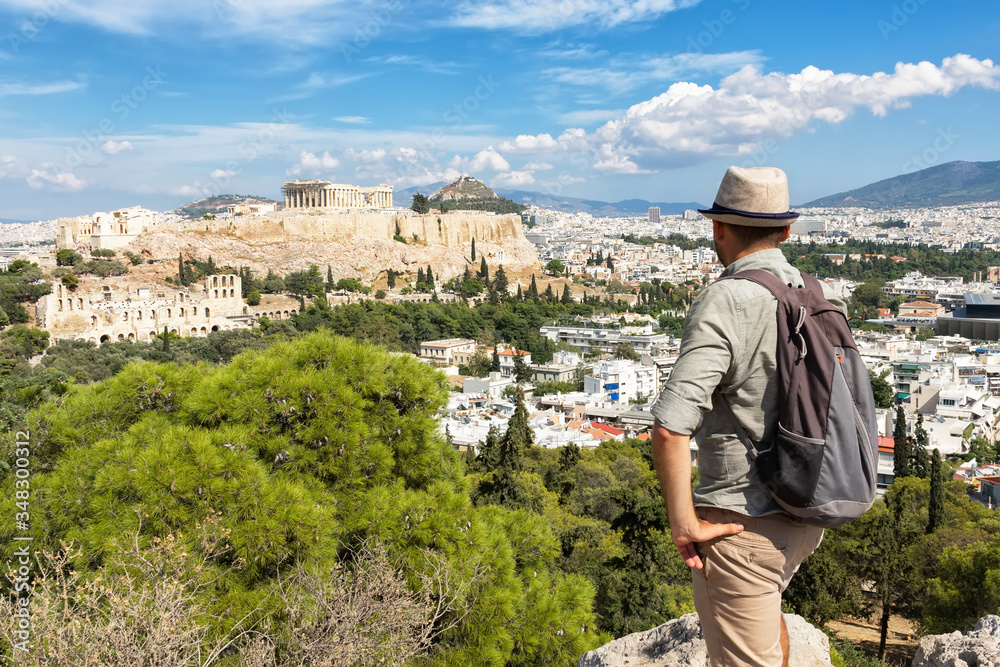 A man in a hat stands on a stone in the center of Athens, in the background the Acropolis and ancient ruins	