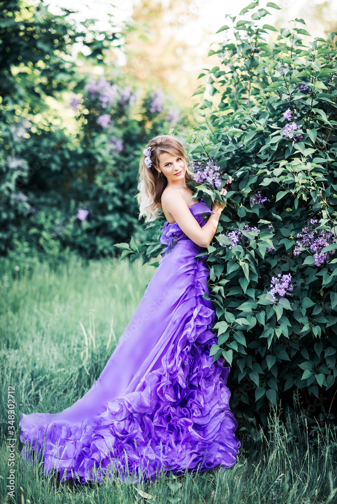 A beautiful young woman enjoys the smell in a blooming spring Lilac garden. Flowers in her hair. She's wearing a long, chic purple dress with a train. Artistic image of high fashion