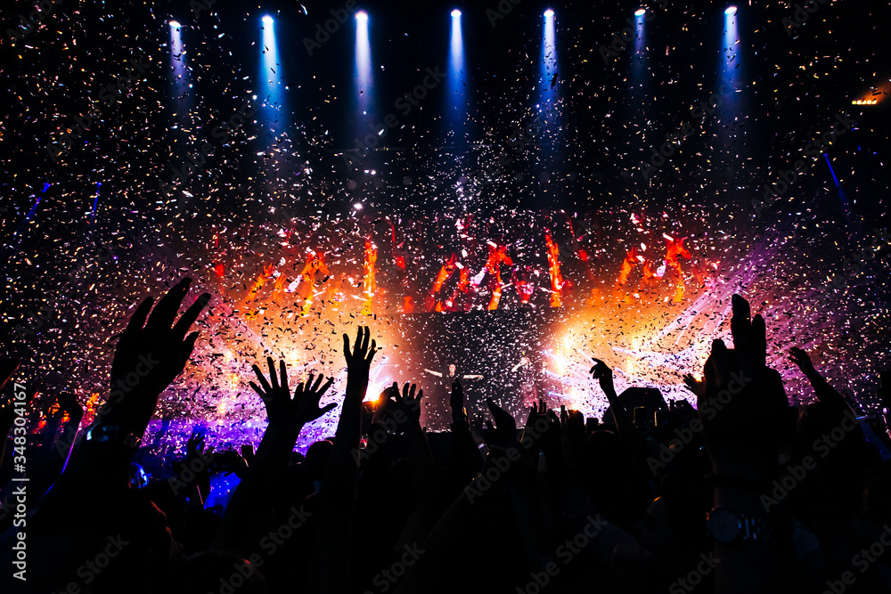 Fans hands raised up during the show. Bright lights and shiny confetti at a pop concert. happy youth dancing at a festival in a crowd.  view of the stage.