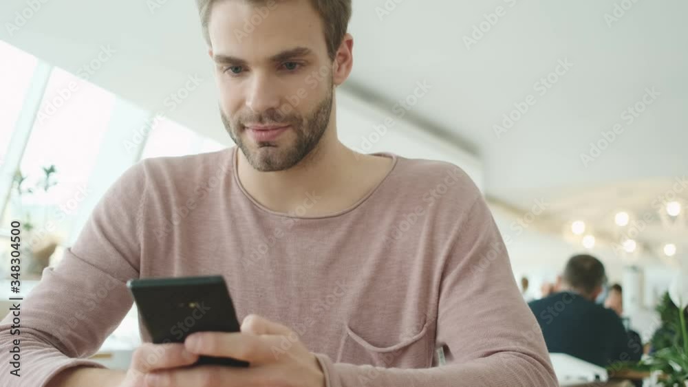 A pleased young bearded man is using his smartphone while sitting in the cafe indoors A pleased young bearded man is using his smartphone while sitting in the cafe indoors