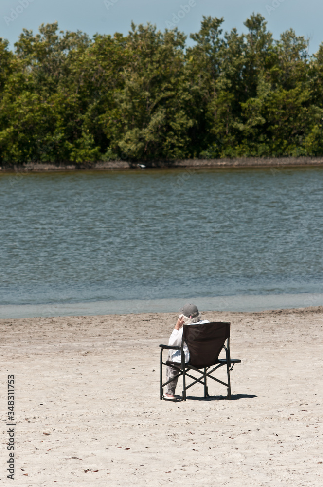 Back view, far distance of a senior woman relaxing in a beach chair on a sandy beach off the coast of an island on gulf of Mexico on a sunny, spring day Back view, far distance of a senior woman relaxing in a beach chair on a sandy beach off the coast of an island on gulf of Mexico on a sunny, spring day