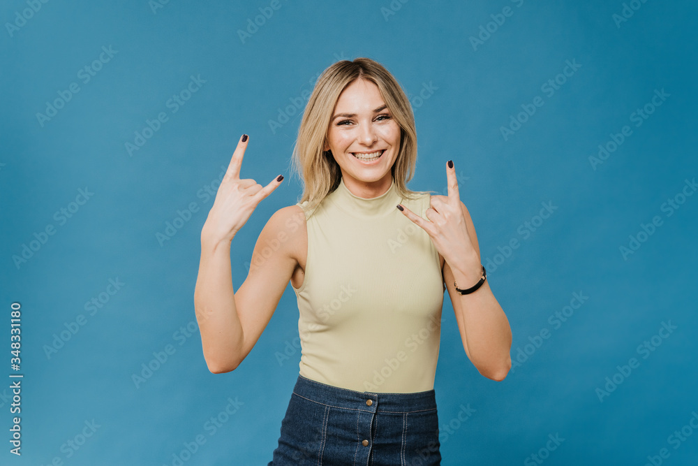 Beautiful positive blonde in a light t-shirt and a denim skirt, shows rock hand gesture broad smiling, against blue backdrop with copy space. People sincere emotions. Beautiful positive blonde in a light t-shirt and a denim skirt, shows rock hand gesture broad smiling, against blue backdrop with copy space. People sincere emotions.