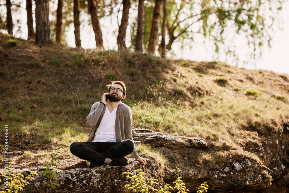 Young bearded hipster man sitting in mountains on a rock, talking on the phone. Office in nature concept.