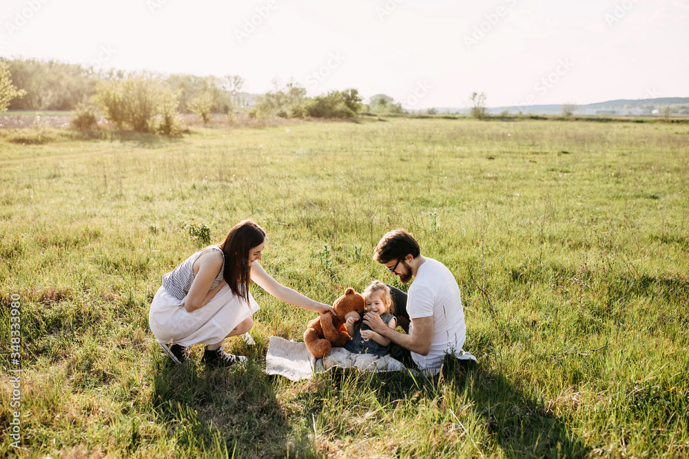 Mom and dad playing with little daughter outdoors, sitting on a blanket in a meadow. Young parents spending time with child. Mom and dad playing with little daughter outdoors, sitting on a blanket in a meadow. Young parents spending time with child.