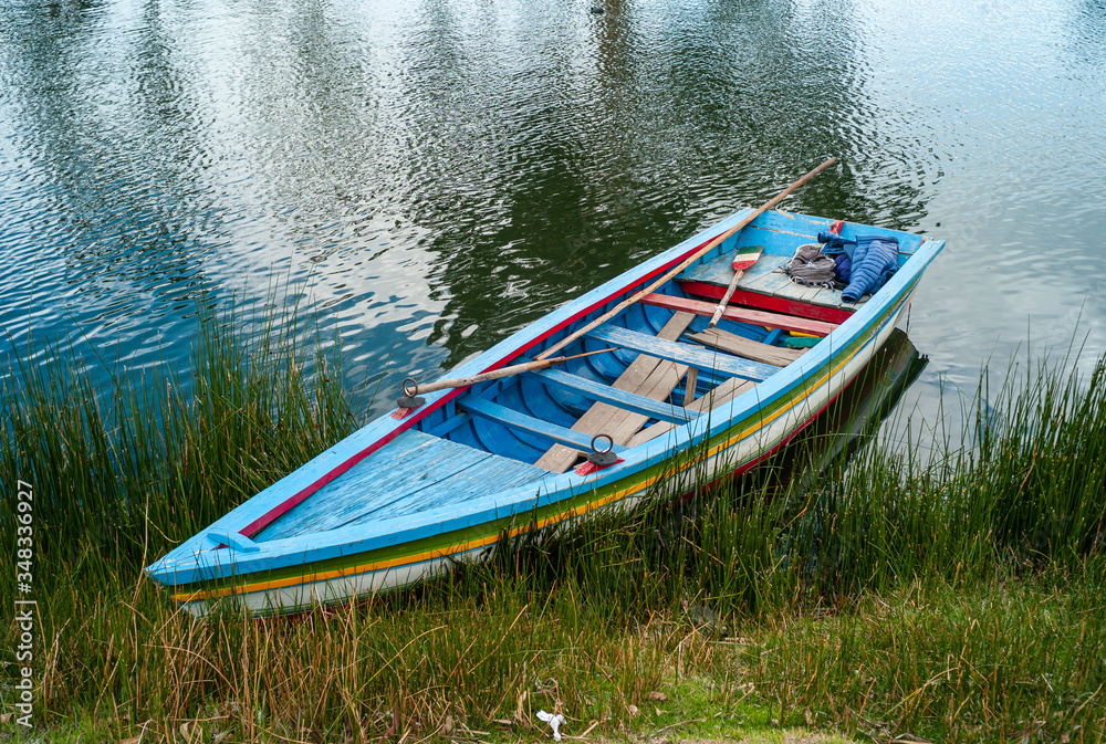 Small Blue, Green, Yellow and White Recreational Boat with Bolivian Flag Themed Paddle by the Touristic Achocalla Lagoon Surrounded by Green Pasture Small Blue, Green, Yellow and White Recreational Boat with Bolivian Flag Themed Paddle by the Touristic Achocalla Lagoon Surrounded by Green Pasture