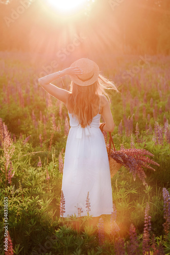 Young blonde girl with long hair in summer dress and a wicker hat with a ribbon. Woman standing with his back at sunset with a large bouquet of lupins.