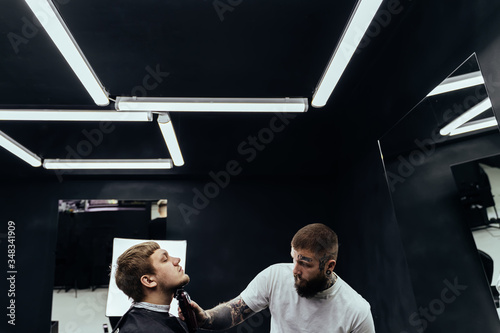 Tattooed Barber trimming bearded man with shaving machine in barbershop. Hairstyling process. Hairstylist cutting the beard of a bearded male.