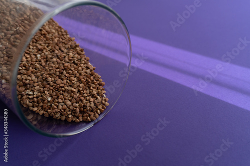 Brown fried buchwheat in a glass jar laying on purple background, cereal concept, close up, copy space, sun light on background. photo