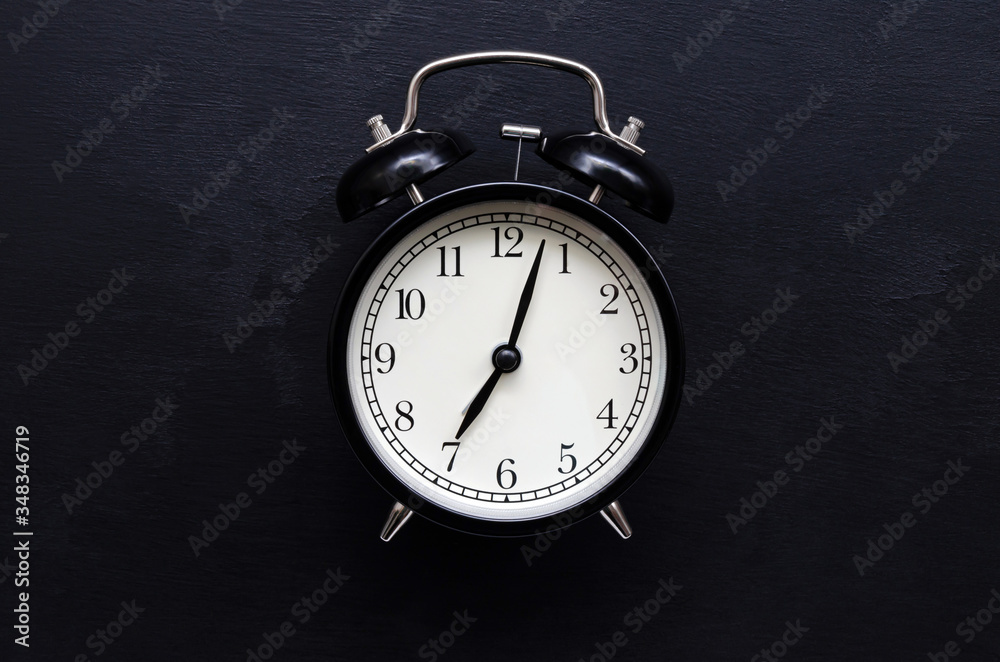 a black alarm clock with a white dial shows the time of 7 hours on a black slate background with place for text a black alarm clock with a white dial shows the time of 7 hours on a black slate background with place for text