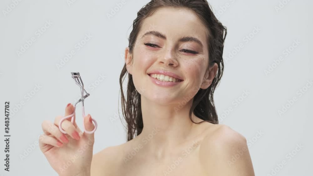 A beautiful young half-naked woman with wet hair is showing a eyelash curler isolated over a light blue background in studio A beautiful young half-naked woman with wet hair is showing a eyelash curler isolated over a light blue background in studio